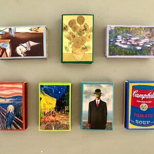 以下が含まれることがあります： 7つのマッチ箱のセットで、それぞれ異なる有名な絵画が描かれています。マッチ箱はすべて同じサイズと形をしていて、一列に並んでいます。絵画には、「叫び」、「星月夜」、「睡蓮」、「キャンベルスープ缶」、「人子の像」、そして他の2つが含まれています。