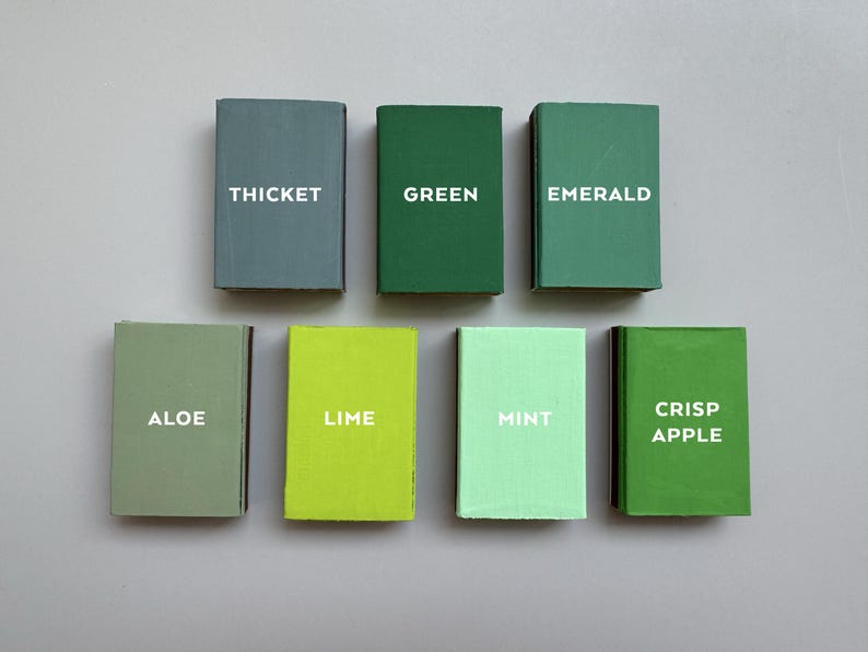 May include: Seven small, rectangular blocks in varying shades of green, each labeled with a different green-related word in white text: Thicket, Green, Emerald, Aloe, Lime, Mint, and Crisp Apple. The blocks are arranged on a gray surface.