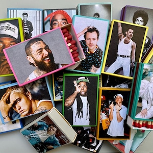 May include: A collection of matchboxes featuring various musicians, including a man with a beard, a man with a crown, a man with a microphone, and a man with a guitar. The matchboxes are arranged in a pile, with some of them overlapping.