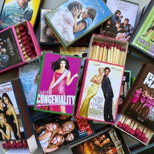 May include: A collection of matchboxes featuring movie posters. The boxes display titles like "Miss Congeniality," "How to Lose a Guy in 10 Days," and "The Notebook." Each box contains wooden matches with red tips, and the boxes are in various colors.