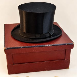 May include: A black silk top hat with a matching ribbon band resting on a square, dark red storage box. Below the box are five bow ties: one black, three off-white, and one cream-colored, each featuring a small metal hook attachment.
