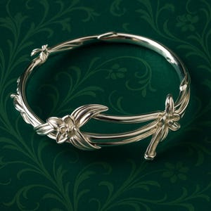 Könnte beinhalten: Ein silberfarbenes Armband mit floralem Design. Das Armband hat stilisierte Blumen und Blätter mit einer glatten, polierten Oberfläche. Das Armband befindet sich vor einem dunkelgrünen Hintergrund mit floralem Muster.