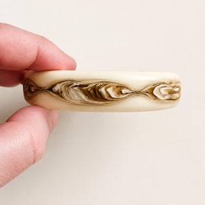 May include: Cream-coloured bangle bracelet with a marbled design in shades of brown and white. The bracelet has a solid, rounded shape, and the marbled pattern creates a flowing, organic aesthetic. A unique piece of jewellery.