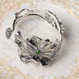 Peut inclure: Un bracelet argenté avec un design floral détaillé, avec un motif de feuille central orné d'une pierre verte. Le design ouvert du bracelet présente des motifs de feuilles complexes et une esthétique délicate inspirée de la nature.