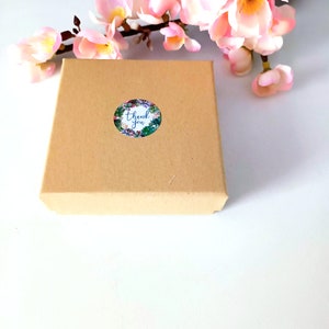 K&ouml;nnte beinhalten: Eine kleine braune Pappschachtel mit einem floralen "Thank You"-Aufkleber. Rosa Bl&uuml;ten sind im Hintergrund. Der Aufkleber hat einen wei&szlig;en Hintergrund mit blauem Text und einem floralen Rand. Die Box ist geschlossen.