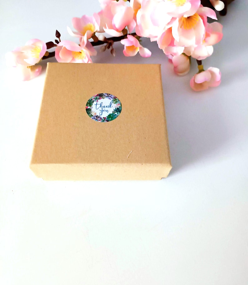 K&ouml;nnte beinhalten: Eine kleine braune Pappschachtel mit einem floralen "Thank You"-Aufkleber. Rosa Bl&uuml;ten sind im Hintergrund. Der Aufkleber hat einen wei&szlig;en Hintergrund mit blauem Text und einem floralen Rand. Die Box ist geschlossen.