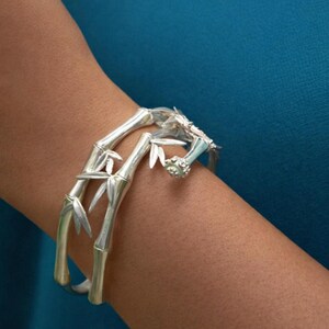 Op de afbeelding: Een zilverkleurige armband in bamboestijl. De armband heeft gedetailleerde bamboestengels en bladeren, waardoor een naturalistisch ontwerp ontstaat. De armband wordt om een arm gedragen, tegen een tealblauwe achtergrond.