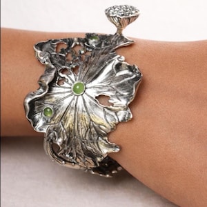 Könnte beinhalten: Silberfarbenes Armband mit Blattmuster und drei grünen Edelsteinen. Das Armband hat ein aufwendiges, strukturiertes Muster und eine kleine, knospenartige Verzierung. Der Schmuck wird am Handgelenk getragen.
