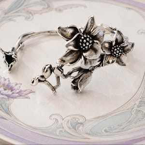 Puede incluir: Un brazalete de plata con un diseño floral detallado que presenta dos flores abiertas, capullos más pequeños y una banda en forma de tallo. El metal tiene un acabado oxidado que resalta los pétalos texturizados y los centros.