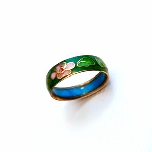 Op de afbeelding: Een vintage ring met een bloemenontwerp. De ring heeft een groene emaille band met roze en witte bloemen en groene bladeren. De binnenkant van de ring is blauw. De ring staat tegen een witte achtergrond.