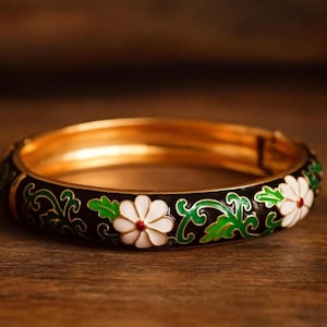 Op de afbeelding: Een goudkleurige armband met een zwarte emaille band. De armband is versierd met witte madeliefachtige bloemen met rode centra en groene bladeren. De achtergrond is een vage houten ondergrond.