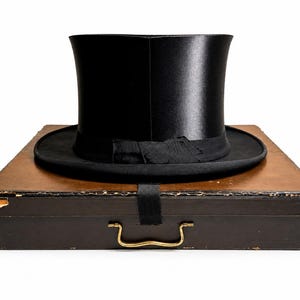 May include: A black top hat with a satin finish, resting on a vintage brown hat box. The hat features a black grosgrain ribbon band. The box has a brass handle and a black strap.