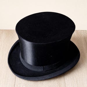 Puede incluir: Un sombrero de copa negro con una corona cilíndrica alta y un ala ancha y curva. El sombrero presenta un acabado suave y satinado en la corona y una cinta de grosgrain negra a juego que rodea la base donde la corona se une al ala.