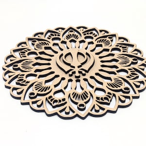 Może przedstawiać: Wycinany laserowo drewniany podkładka pod szklankę o skomplikowanym, symetrycznym wzorze. Podkładka ma centralny emblemat i szczegółowy, ażurowy wzór w odcieniach brązu i czerni. Średnica wynosi około 10 cm.