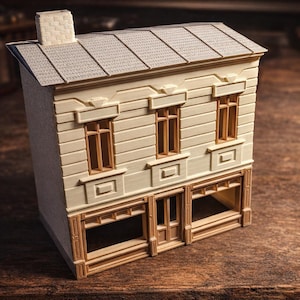 Könnte beinhalten: Ein Miniaturmodell eines zweistöckigen Gebäudes mit hellbeiger Fassade und grauem Ziegeldach. Das Gebäude verfügt über detaillierte Fensterrahmen, einen Schornstein und offene Schaufenster im Erdgeschoss. Das Modell steht auf einer Holzoberfläche.
