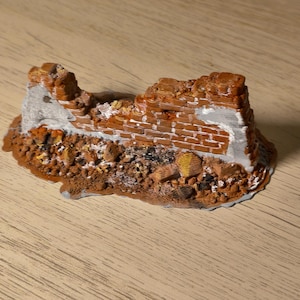 May include: A miniature diorama piece depicting a ruined brick wall section. The structure features weathered red bricks with patches of grey mortar and exposed concrete. The base is covered in textured brown rubble, debris, and scattered stone fragments.