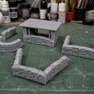 May include: A set of gray miniature sandbag fortifications including a small rectangular bunker with a flat roof and three separate wall sections of varying shapes, all crafted with detailed stacked sandbag textures for tabletop gaming.