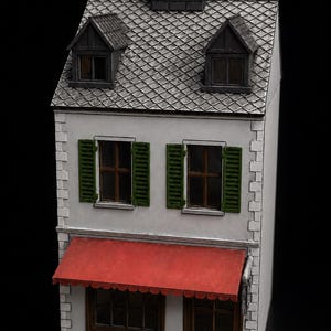 Puede incluir: Una maqueta en miniatura de un edificio de dos plantas con tejado de tejas grises, chimenea central y dos buhardillas. La fachada es blanca con contraventanas verdes en las ventanas superiores, un toldo rojo y una puerta marrón.