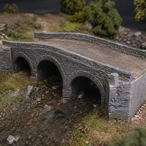 Op de afbeelding: Een miniatuurmodel van een grijze stenen brug met drie boogopeningen. De brug heeft een getextureerd metselwerkoppervlak met lage stenen muren aan de zijkanten, die over een kleine beek met rotsachtige oevers en stukjes groen gras loopt.