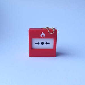 Può includere: Una piccola scatola rossa di allarme antincendio con un pannello bianco con un'icona a forma di fiamma e frecce direzionali. Ha una catena dorata attaccata. Il design è semplice e grafico, con un'estetica pulita e moderna.