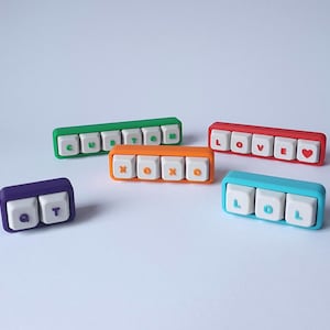 Può includere: Una collezione di oggetti colorati a forma di tasti della tastiera. I tasti sono di vari colori, tra cui verde, arancione, rosso, blu e viola. Ogni tasto ha una parola o lettere diverse, come "CUSTOM", "XOXO", "LOVE", "LOL" e "QT".