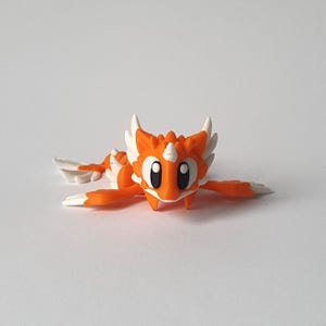Puede incluir: Una pequeña figura de dragón naranja y blanco con grandes ojos negros, un cuerno blanco y estructuras blancas en forma de alas. El dragón está tumbado sobre una superficie blanca, con los brazos y las alas extendidas.