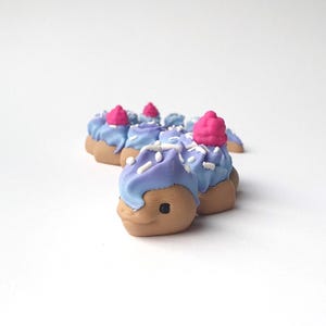 Op de afbeelding: Een verzameling kleine, cupcake-vormige beeldjes. Elk beeldje heeft een bruine basis, blauwe frosting met een paarse swirl, een roze kers en witte hagelslag. De beeldjes zijn in een halve cirkel gerangschikt.