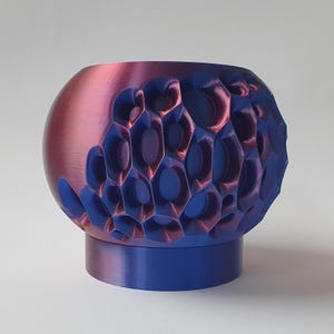 Pode incluir: Objeto decorativo esférico com um padrão texturizado em forma de favo de mel. A parte superior é um gradiente de vermelho a roxo, enquanto a seção estampada e a base são azuis. O objeto é provavelmente impresso em 3D.