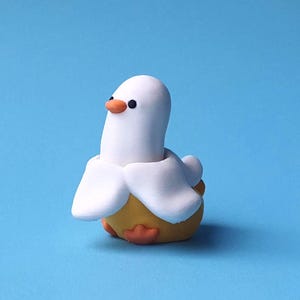 Könnte beinhalten: Eine skurrile, handgefertigte Figur, die eine Ente-Banane-Hybride darstellt. Die Ente hat einen weißen Kopf und Flügel, einen orangefarbenen Schnabel und Füße sowie einen gelben, bananenförmigen Körper. Der Hintergrund ist einfarbig hellblau.