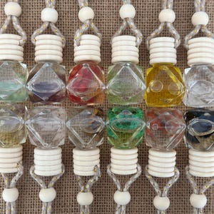 May include: Twelve clear glass bottles with various colored liquids inside. Each bottle has a white wooden cap and is attached to a string with a white bead. The bottles are hanging from a brown burlap background.