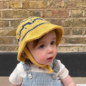 May include: A baby wearing a yellow crocheted sun hat with blue trim and a blue and white striped overall.
