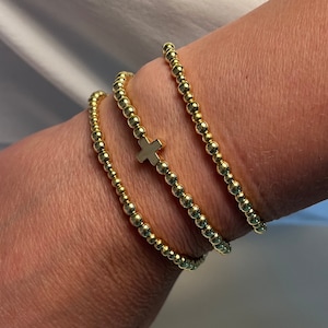 Może przedstawiać: Trzy złote bransoletki z koralików. Jedna bransoletka ma mały złoty krzyżyk. Bransoletki wykonane są z małych, okrągłych, złotych koralików. Bransoletki są ułożone jedna na drugiej.