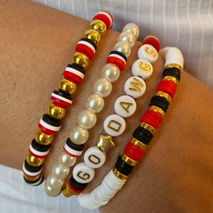 May include: Four bracelets stacked on a wrist. The top bracelet is black, white, and red beads with gold accents. The second bracelet is white pearl beads. The third bracelet has white beads with the letters "GO DAWGS" in gold. The bottom bracelet is black, white, and red beads with gold accents.
