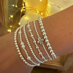 May include: A collection of silver beaded bracelets, some with small white pearls and a cross charm. The bracelets are worn on a wrist, showcasing their delicate design and shine. The bracelets are a mix of different styles.