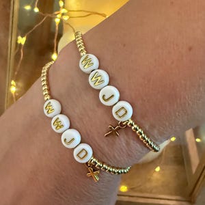 Puede incluir: Dos pulseras de oro con cuentas blancas que deletrean "WJD" con un pequeño dije de cruz dorada en cada pulsera.