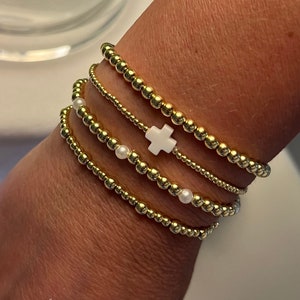 May include: A set of four gold beaded bracelets with white pearl accents. One bracelet features a white cross charm.