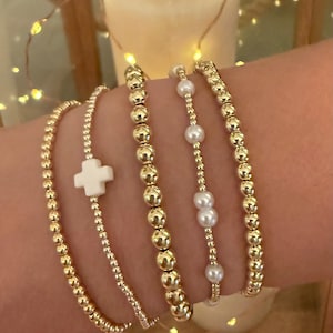 May include: A set of five stretch bracelets featuring metallic gold-toned beads. One bracelet includes a white cross charm, while another alternates between small gold-toned beads and round white pearls. The remaining three bracelets consist of uniform gold-toned beads.