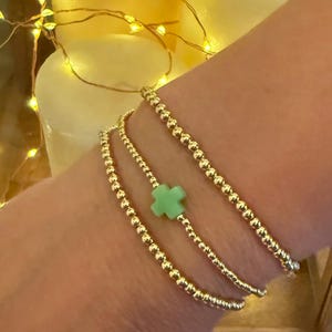 May include: Three gold beaded bracelets stacked on a wrist. The top bracelet has a light green cross charm.