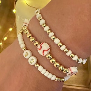 May include: Three white beaded bracelets with gold beads and a baseball charm. One bracelet has the number "22" on two white beads.