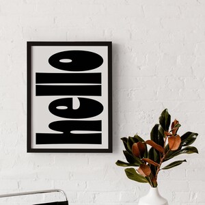 May include: Black and white framed print with the word "hello" in bold, block letters. The print is hanging on a white brick wall, with a black leather chair and a white vase with a green plant in the foreground.