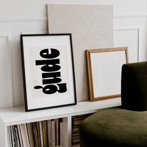 Puede incluir: Una impresión artística enmarcada con la palabra "guide" en letras negras y en negrita. La impresión se exhibe en una estantería blanca con libros, junto a una impresión con marco dorado y un lienzo texturizado. Una silla de terciopelo verde está en primer plano.