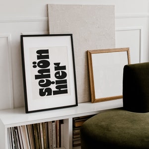 Puede incluir: Una estantería blanca con una impresión enmarcada en negro que dice "schön hier" en letras negras en negrita. Una imagen enmarcada en dorado está a la derecha de la impresión. Un sillón de terciopelo verde oscuro está en primer plano.