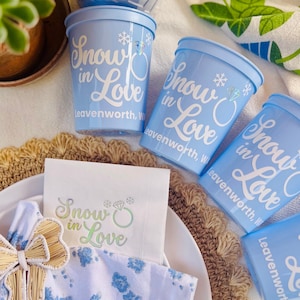 Puede incluir: Vasos de plástico azul claro y una servilleta con el texto "Snow in Love" y un gráfico de anillo de diamantes. Los vasos están dispuestos con un lazo decorativo y una planta en el fondo. Para una boda o compromiso.