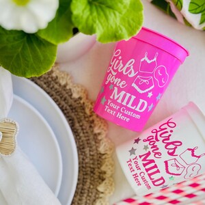 May include: Two plastic cups, one pink and one white, with the text "Girls Gone Mild" and custom text. The cups feature illustrations of lingerie and stars. The cups are on a white surface with a decorative placemat and plates.