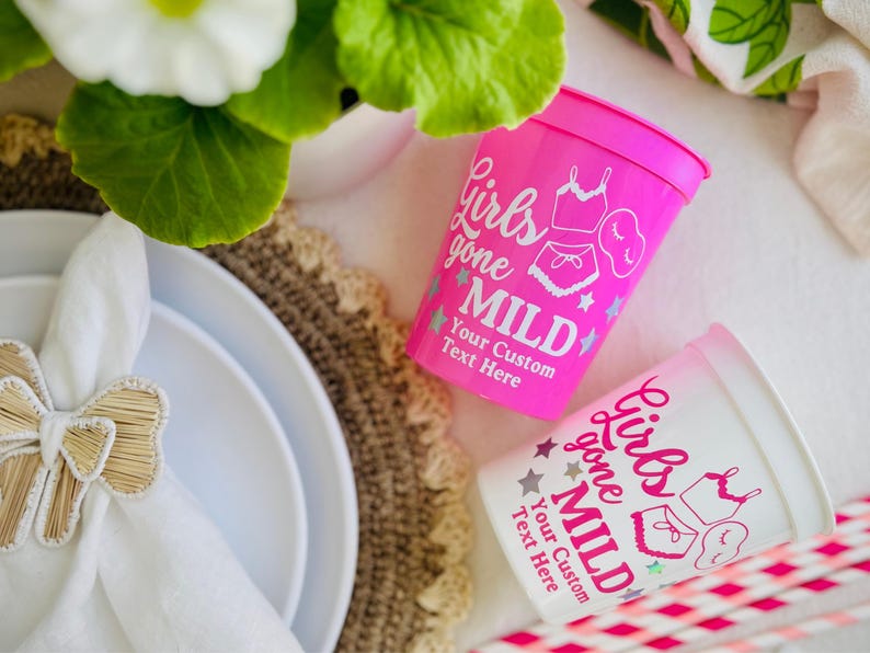 May include: Two plastic cups, one pink and one white, with the text "Girls Gone Mild" and custom text. The cups feature illustrations of lingerie and stars. The cups are on a white surface with a decorative placemat and plates.