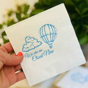 Op de afbeelding: Witte papieren servet met een blauwe illustratie van een luchtballon en een wolk. De tekst "We are on Cloud Nine" is in blauw gedrukt.