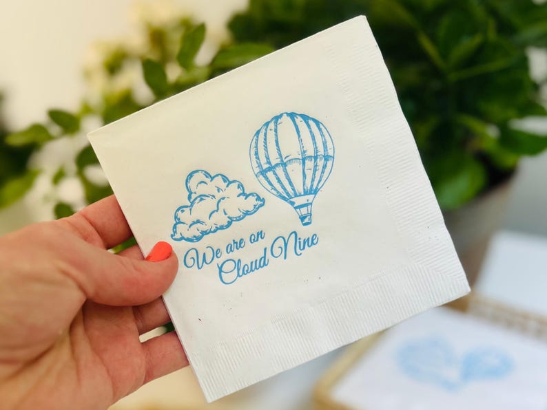 Op de afbeelding: Witte papieren servet met een blauwe illustratie van een luchtballon en een wolk. De tekst "We are on Cloud Nine" is in blauw gedrukt.