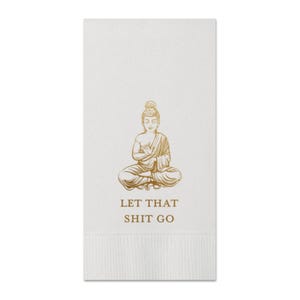 May include: A white paper napkin with a gold illustration of a seated Buddha and the text "LET THAT SHIT GO". The Buddha is in a meditative pose, and the text is in a simple, sans-serif font. The napkin has a textured bottom.