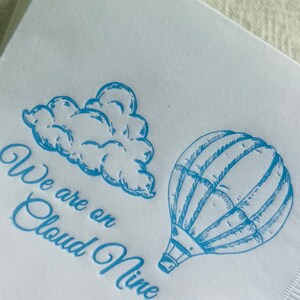 Op de afbeelding: Een witte servet met blauwe inkt erop gedrukt. De servet heeft een tekening van een luchtballon en een wolk. De tekst luidt "We are on Cloud Nine".