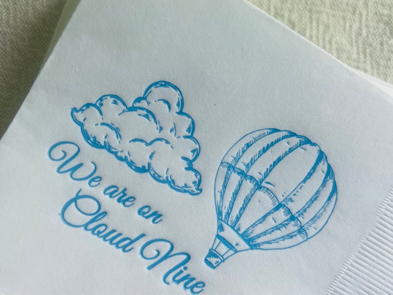 Op de afbeelding: Een witte servet met blauwe inkt erop gedrukt. De servet heeft een tekening van een luchtballon en een wolk. De tekst luidt "We are on Cloud Nine".